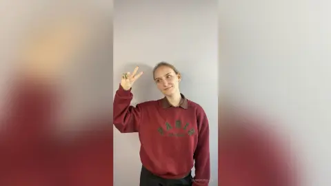 Smiling young woman in a burgundy oversized sweatshirt posing with a peace sign against a plain wall.
