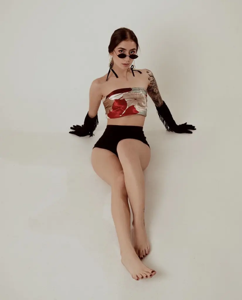 scarletjonessX poses with a direct gaze, wearing dark sunglasses and black gloves with a patterned, light-colored top against a plain background.