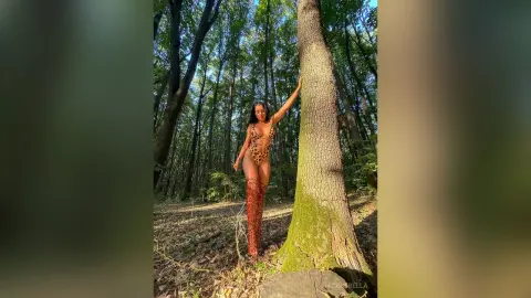 NicoleBella stands naked in a forest setting, wearing a leopard print bodysuit and tall red boots while leaning against a large tree trunk.