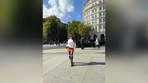 Stylish woman in a white sweater and orange skirt walking on a sunny city street near historic buildings.