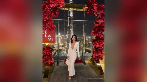 Elegant woman in a white evening gown posing by a floral archway with a city skyline backdrop at night.