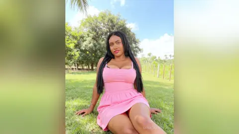 Smiling woman in a pink summer dress sitting on a lush green lawn in a sunny outdoor garden setting.