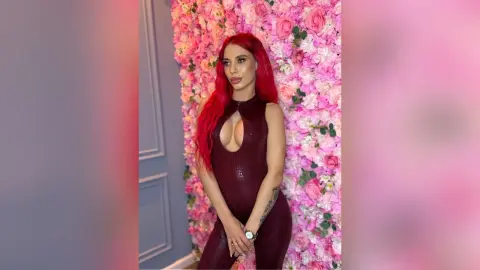 Red-haired woman in a sleek burgundy bodysuit posing against a romantic pink floral wall background.
