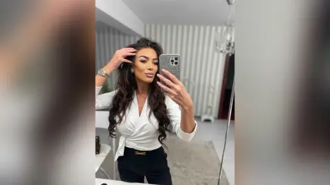 ArianaKroft poses for a mirror selfie wearing a white long-sleeve blouse and black pants, showing off her long wavy dark hair.