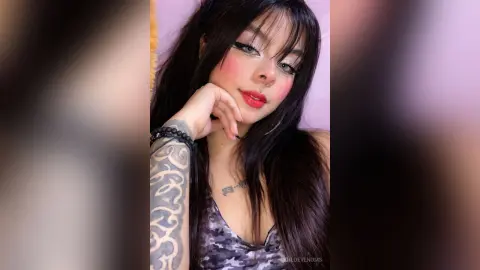 Young woman with long dark hair and tattoos wearing a camouflaged top against a soft pink background.