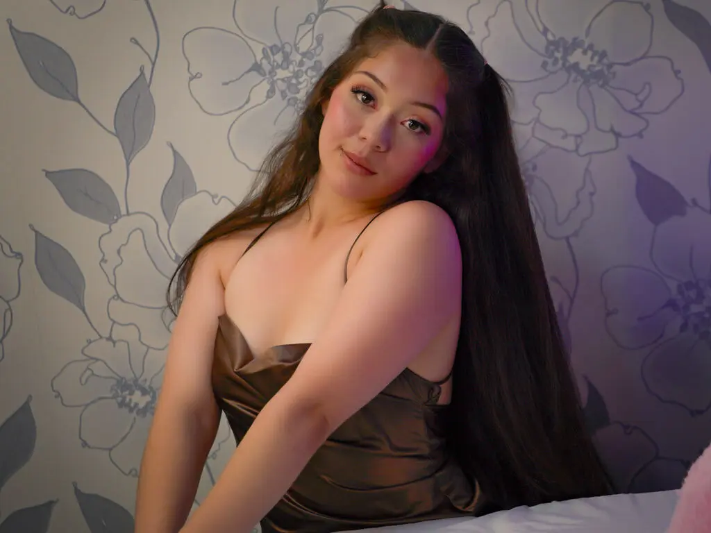 KateAldana poses with long dark hair wearing a brown satin camisole against a floral wallpaper background.