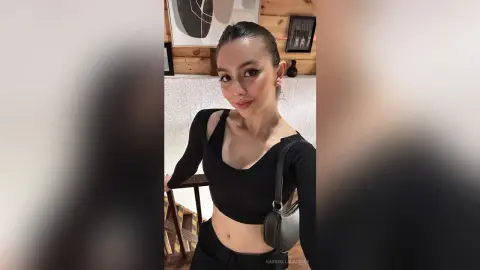Smiling woman in a black long-sleeve crop top and crossbody bag posing in a cozy wood-paneled room.