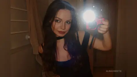 Young woman with long dark hair in a black tank top taking a moody selfie in a dimly lit room.