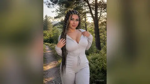 Smiling woman with long braids in a grey lounge jumpsuit standing on a forest path during golden hour.