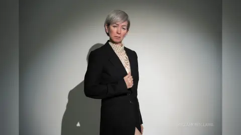 Elegant woman in a black blazer and white lace turtleneck posing against a minimalist grey studio background.