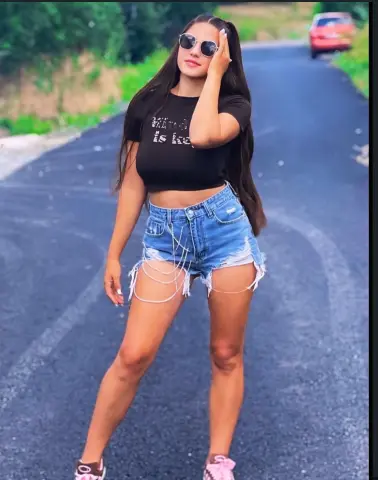 Young woman in black t-shirt and denim shorts posing on a sunny road with a relaxed summer vibe.