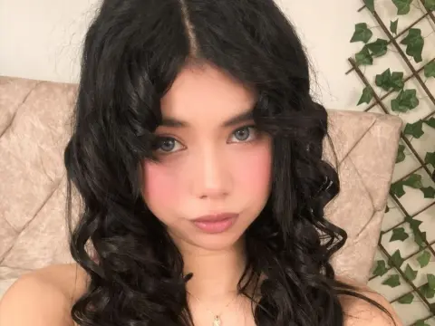 Nathy_Pelaez poses with long, wavy black hair and pink blush against a tufted headboard.