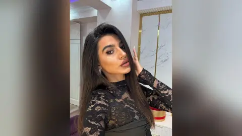 Glamorous woman in a black lace long-sleeve top posing in a modern, bright interior setting.