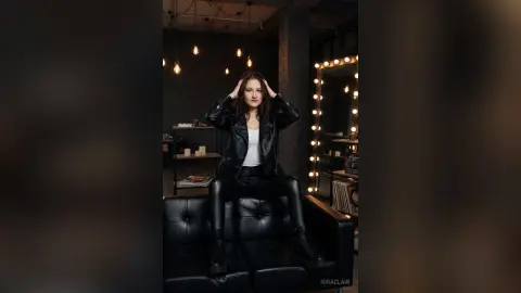 Stylish woman in a black leather jacket and jeans sitting on a sofa in a moody, modern loft setting.