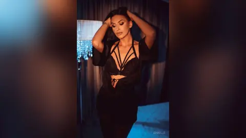 Iza — Elegant woman in a black lace bodysuit and sheer cover-up posing in moody, dim indoor lighting.