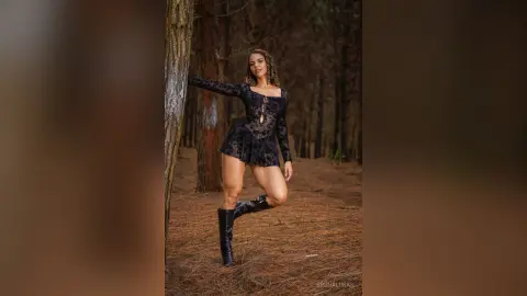 Elegant woman in a black lace bodysuit and tall boots posing in a moody, sunlit forest setting.