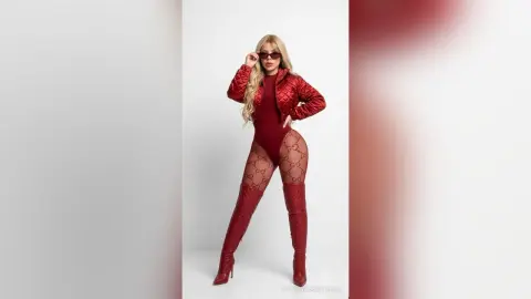 NatashaCastello poses in a red bodysuit, red jacket, and tall red boots against a white background.