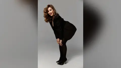 Smiling woman in a chic black blazer and tights posing joyfully against a minimalist grey studio background.