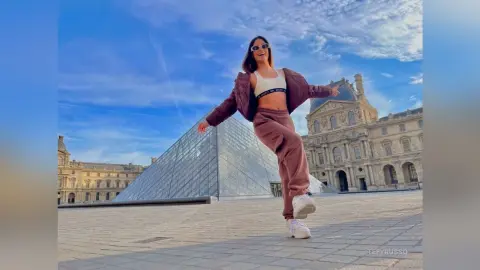 TefyRusso smiles in a white sports bra and matching brown sweatpants, posing outside the Louvre pyramid in Paris.