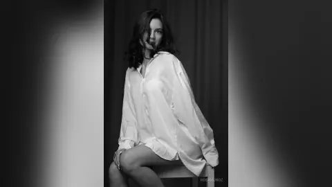 Elegant woman in an oversized white button-down shirt posing in a moody black and white studio portrait.