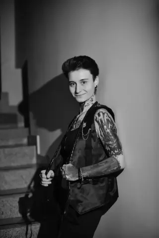Smiling tattooed woman in a dark vest standing on indoor stairs in a moody black and white portrait.
