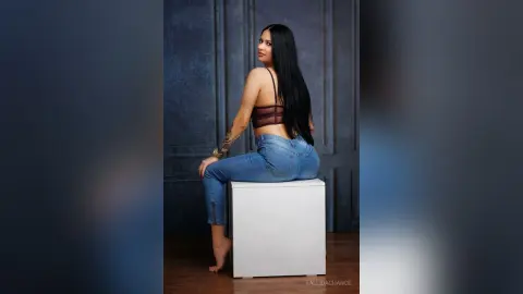 Young woman in denim jeans and burgundy crop top sitting on a white cube against a dark blue studio background.