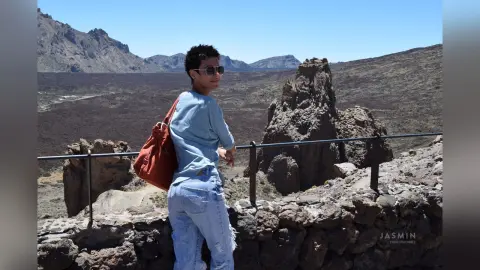 Young man in a blue shirt and jeans standing by a stone wall in a sunny, rocky volcanic landscape.