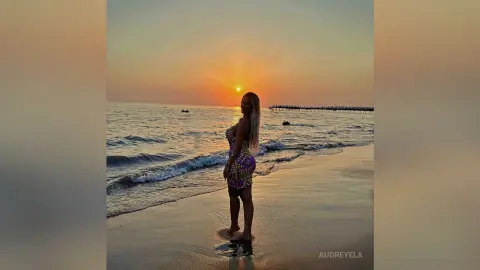 Blonde woman in a floral mini dress standing on a beach at sunset with ocean waves.