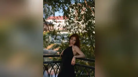 Redheaded woman in a black dress leaning on a balcony railing amidst lush green trees in soft natural light.