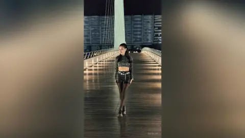 Stylish woman in a black glittery top and leather shorts standing on a modern pedestrian bridge at night.