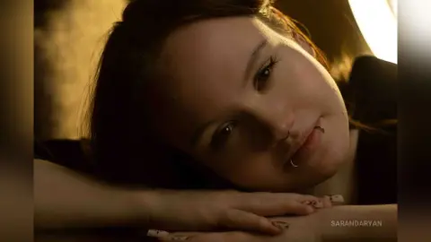 Young woman with nose rings leaning on a table in warm, moody indoor lighting.