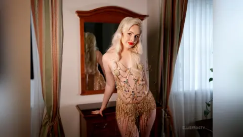 EllieFrosty poses in a gold beaded fringe bodysuit, standing next to a wooden mirror in a room with long curtains.