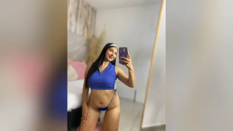 Smiling woman in a blue sleeveless crop top and matching bikini bottoms taking a mirror selfie in a bright room.