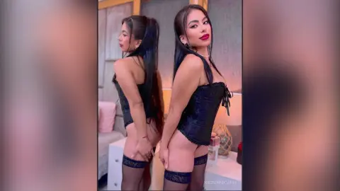 VictoriaCapri poses in a black lace corset and black stockings, looking over her shoulder with long dark hair and red lipstick.