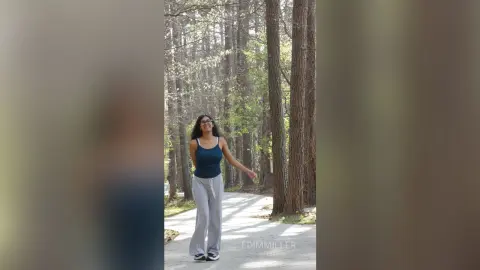 Smiling woman in a navy tank top and grey sweatpants walking through a sunny, peaceful forest path.