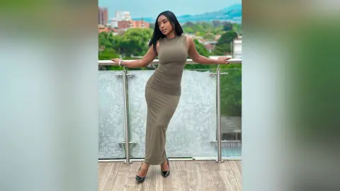 Elegant woman in a sleek olive maxi dress posing on a balcony overlooking a lush green cityscape.