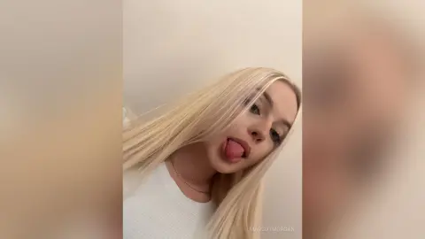Blonde woman in a white top making a playful expression against a neutral background.