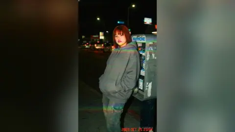Young woman in a grey oversized hoodie standing on a city street at night with a nostalgic vintage mood.