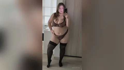 Curvy woman in brown lace lingerie and black thigh-high stockings posing for a mirror selfie in a bright room.