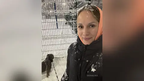 Smiling woman in a black winter jacket and peach hoodie standing in a snowy outdoor setting.