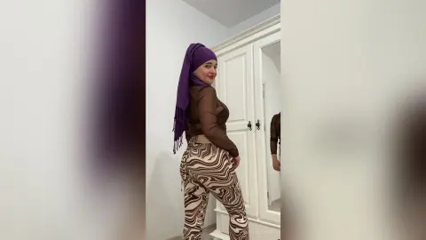 Smiling woman in a purple hijab and brown top wearing patterned trousers in a bright modern room.