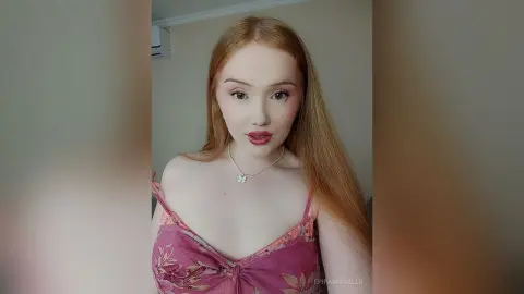 Redheaded woman in a pink floral lace camisole and silver necklace posing with soft makeup in a bright room.
