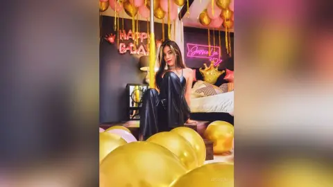 JessieLiu poses in black leather pants and a black bra, surrounded by gold balloons in a room with a pink neon sign.