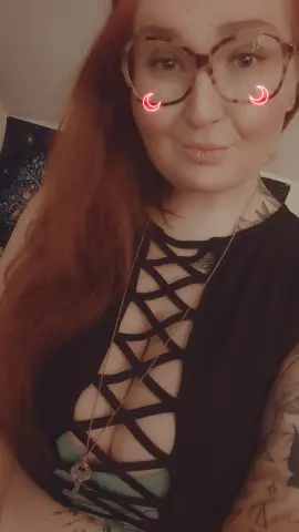 Smiling redhead woman with glasses wearing a black cutout top and silver necklace in a cozy indoor setting.