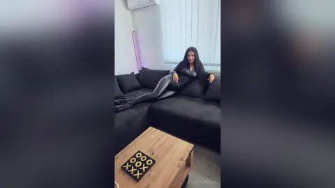 Glamorous woman in a black bodysuit and leather pants relaxing on a dark sofa in a modern living room.