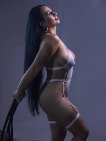 ValentinaJaramilloo poses in a white cut-out bodysuit, showing her long black hair and curves against a plain grey background.