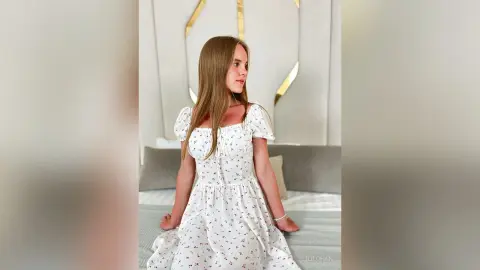 LiliLohan sits on a bed wearing a white floral print sundress with short puffed sleeves.
