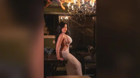 DayanaHille poses in a white cut-out bodysuit, looking over her shoulder in a dimly lit room with a chandelier.