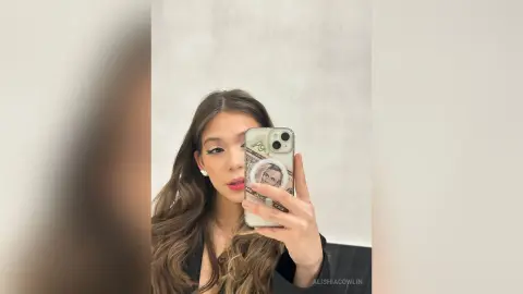 Young woman with long wavy hair taking a selfie in a mirror wearing a black outfit in a bright room.