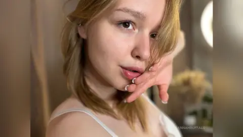 Blonde woman in a white tank top posing with a soft, dreamy expression in warm indoor lighting.